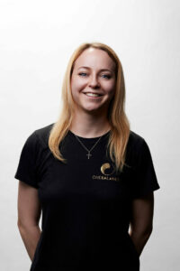 Studio-Porträt einer lächelnden Frau mit blondem Haar im schwarzen T-Shirt vor hellem Hintergrund.