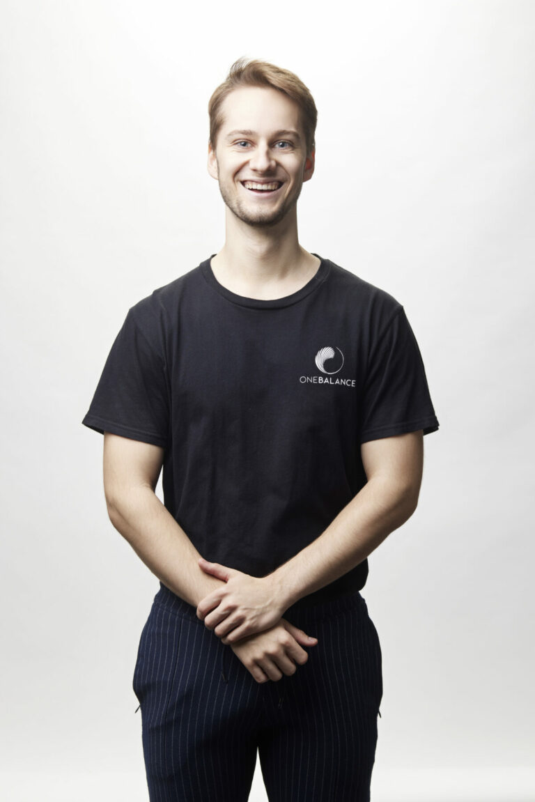 Lächelnder Mann im schwarzen T-Shirt steht vor weißem Hintergrund und hält die Hände locker vor dem Körper.