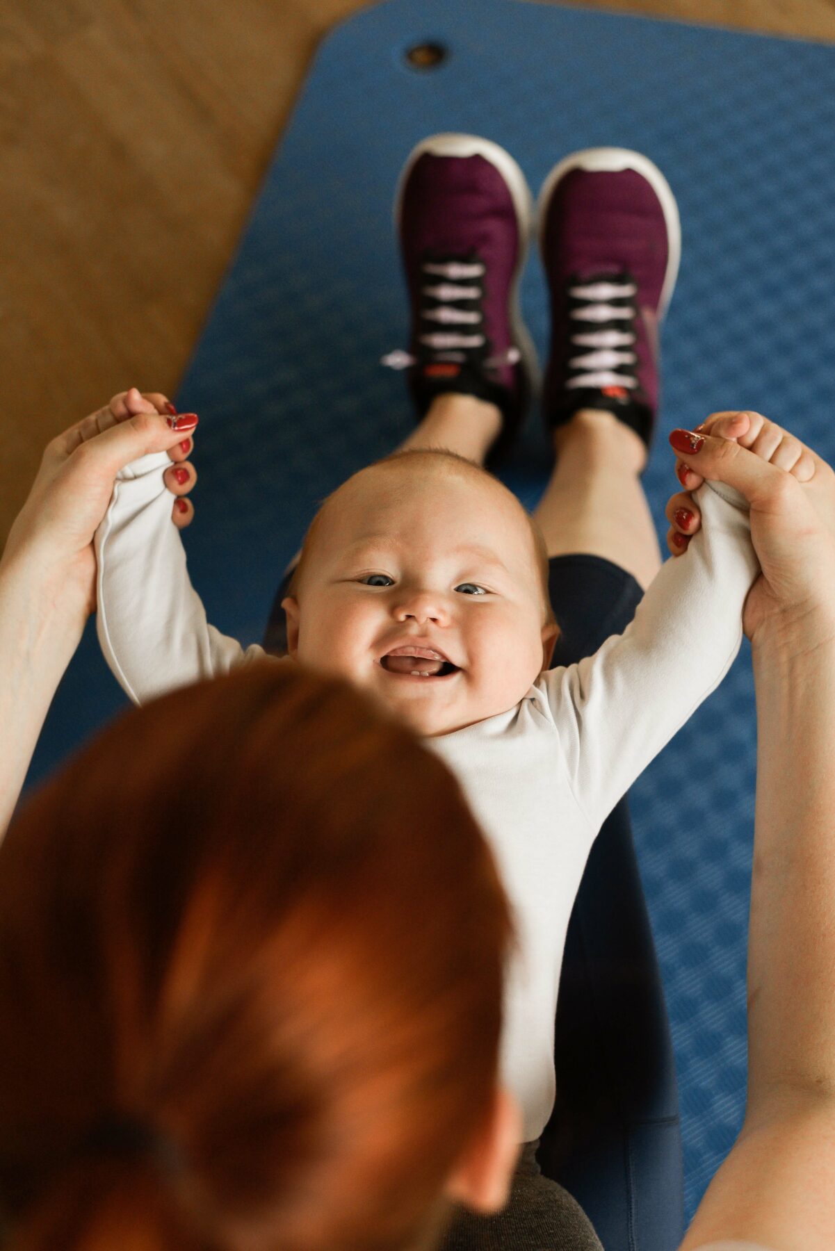 Lachendes Baby hält die Hände eines Erwachsenen, während es auf einer blauen Fitnessmatte liegt.