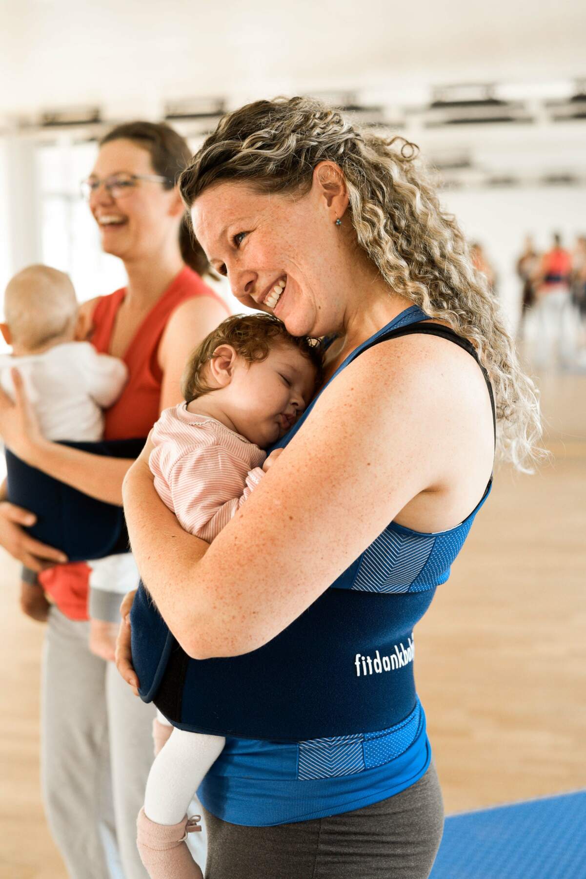 Lächelnde Mutter trägt ein schlafendes Baby in einer Bauchtrage während eines Kurses im hellen Fitnessstudio.