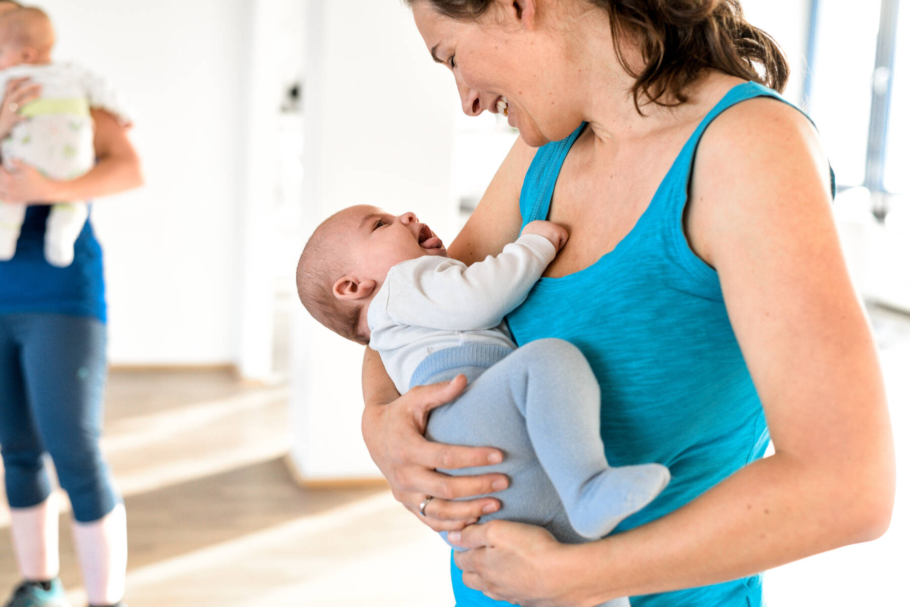 Eine Mutter hält ihr lachendes Baby im Arm und schaut es liebevoll an, im Hintergrund steht eine weitere Person mit Kind.