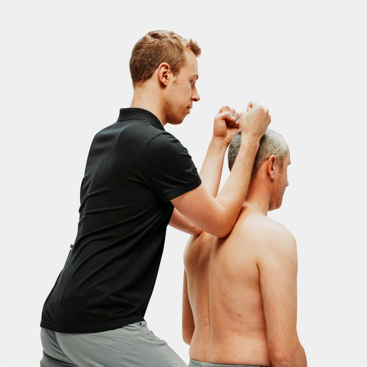 Physiotherapeut führt manuelle Therapie am Nacken und an den Schultern eines Patienten durch, Seitenansicht vor weißem Hintergrund.